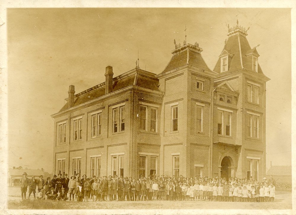Sepia photograph of the early multistory Lyons High School building, with a large group gathered along its base.