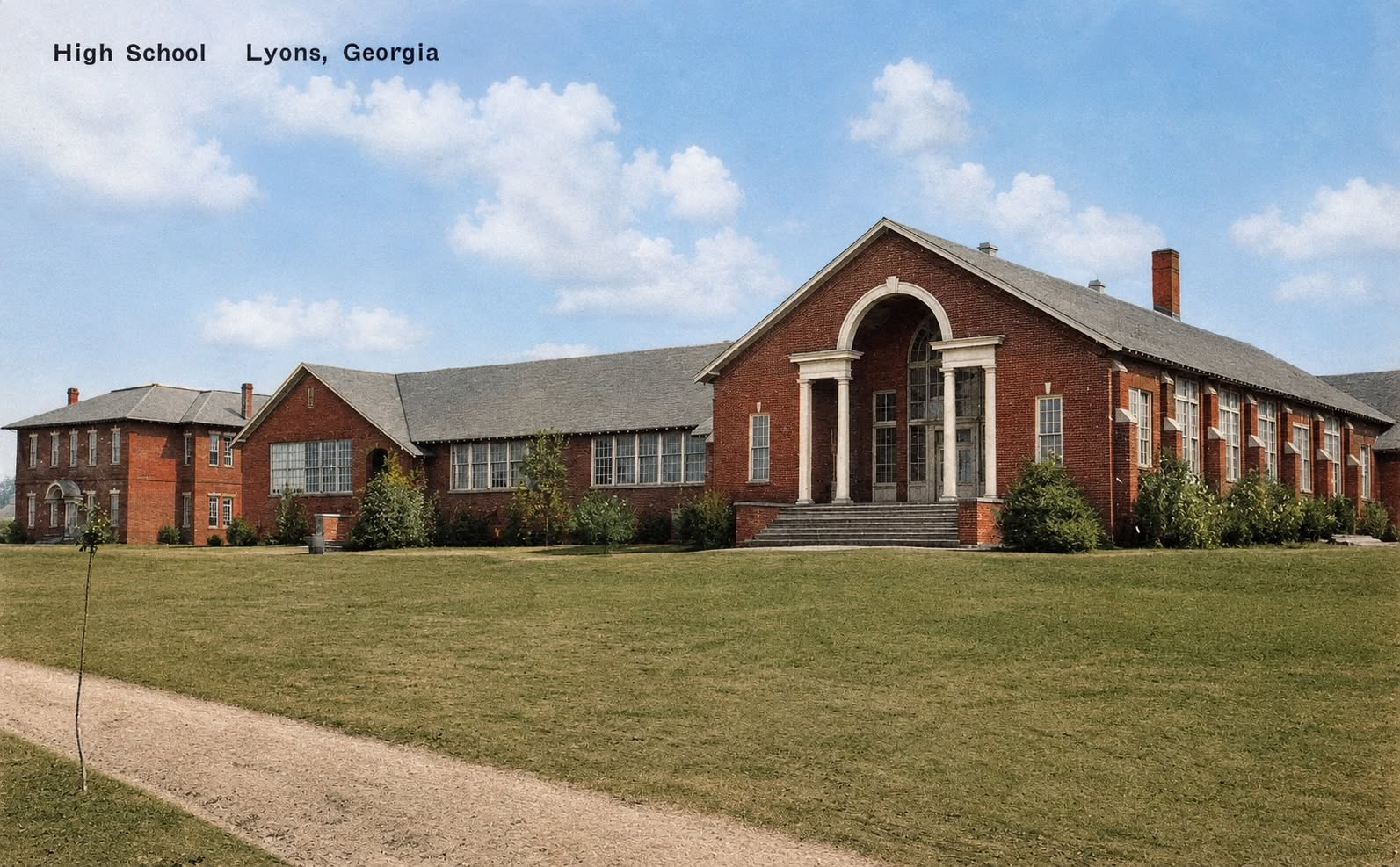 Lyons High School before the fire: a long brick school building with a columned central entrance, green lawn, and blue sky.